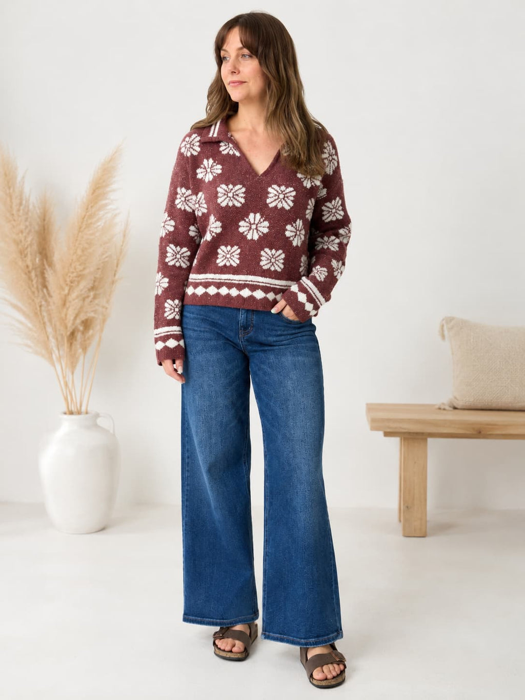 A woman modeling a burgundy knit sweater with a white Nordic snowflake pattern, paired with wide-leg blue jeans and brown sandals, standing in a bright room with a pampas grass vase and a wooden bench.