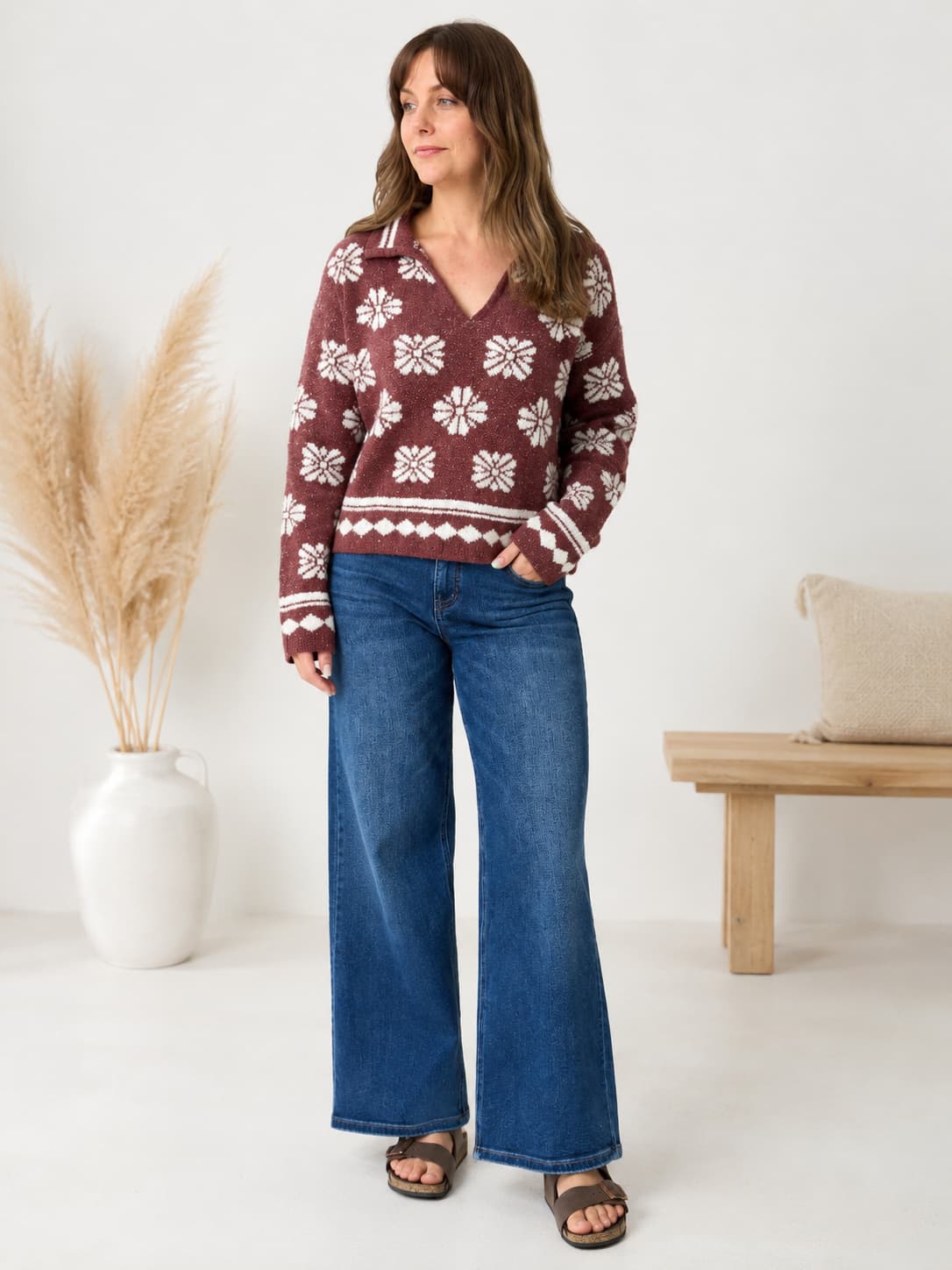 A woman modeling a burgundy knit sweater with a white Nordic snowflake pattern, paired with wide-leg blue jeans and brown sandals, standing in a bright room with a pampas grass vase and a wooden bench.