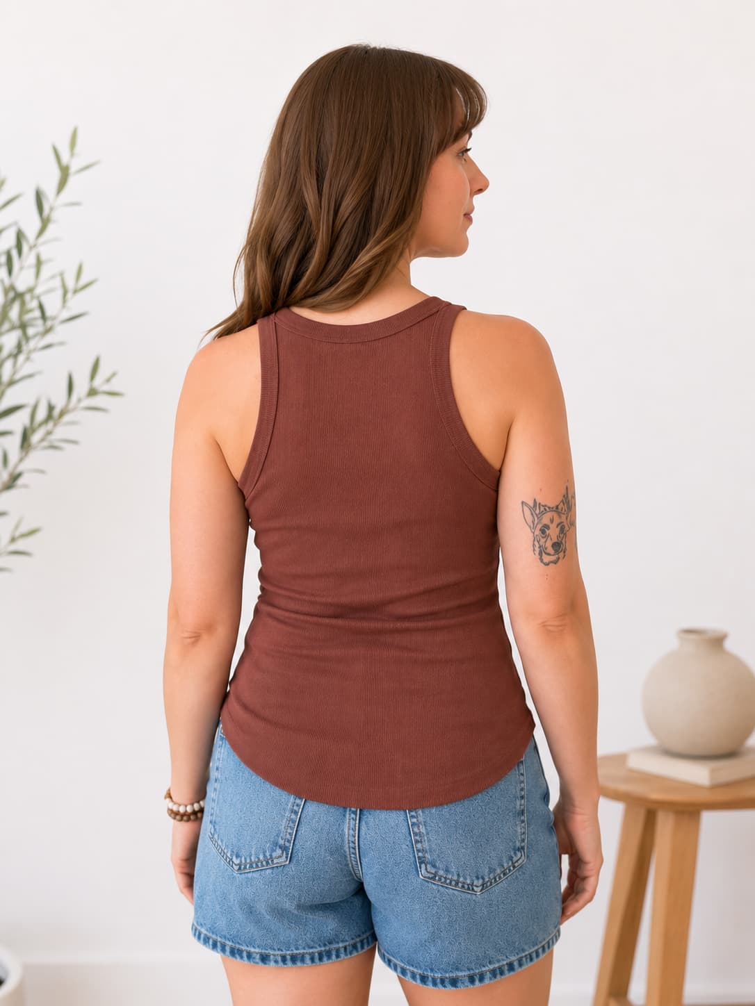 A woman shown from the back wearing a fitted sleeveless tank top in a muted terracotta brown color, paired with light blue denim shorts, standing in a bright indoor setting.