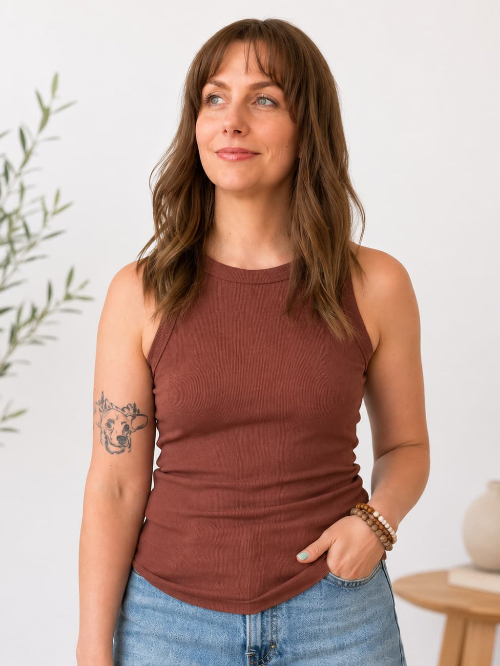A woman wearing a sleeveless rust-brown tank top and light blue jeans, standing against a white wall with a small plant and wooden side table in the background.