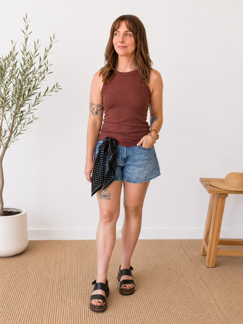 A woman modeling a sleeveless rust-brown tank top, denim shorts, and black sandals in a bright room with a potted plant and wooden stool.