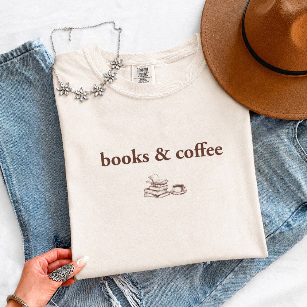 A folded cream-colored t-shirt with 'books & coffee' printed in brown text and a small book-and-cup graphic, styled on top of blue jeans with a brown hat and a silver flower necklace nearby. A hand is visible holding the folded shirt.