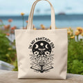Beige tote bag with black graphic design featuring text and symbols, placed outdoors with flowers and water in the background.
