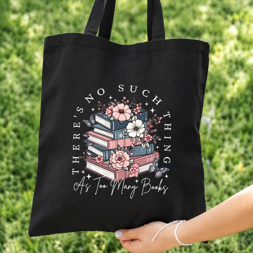 Black tote bag with floral book design and text held by a person against a green grass background