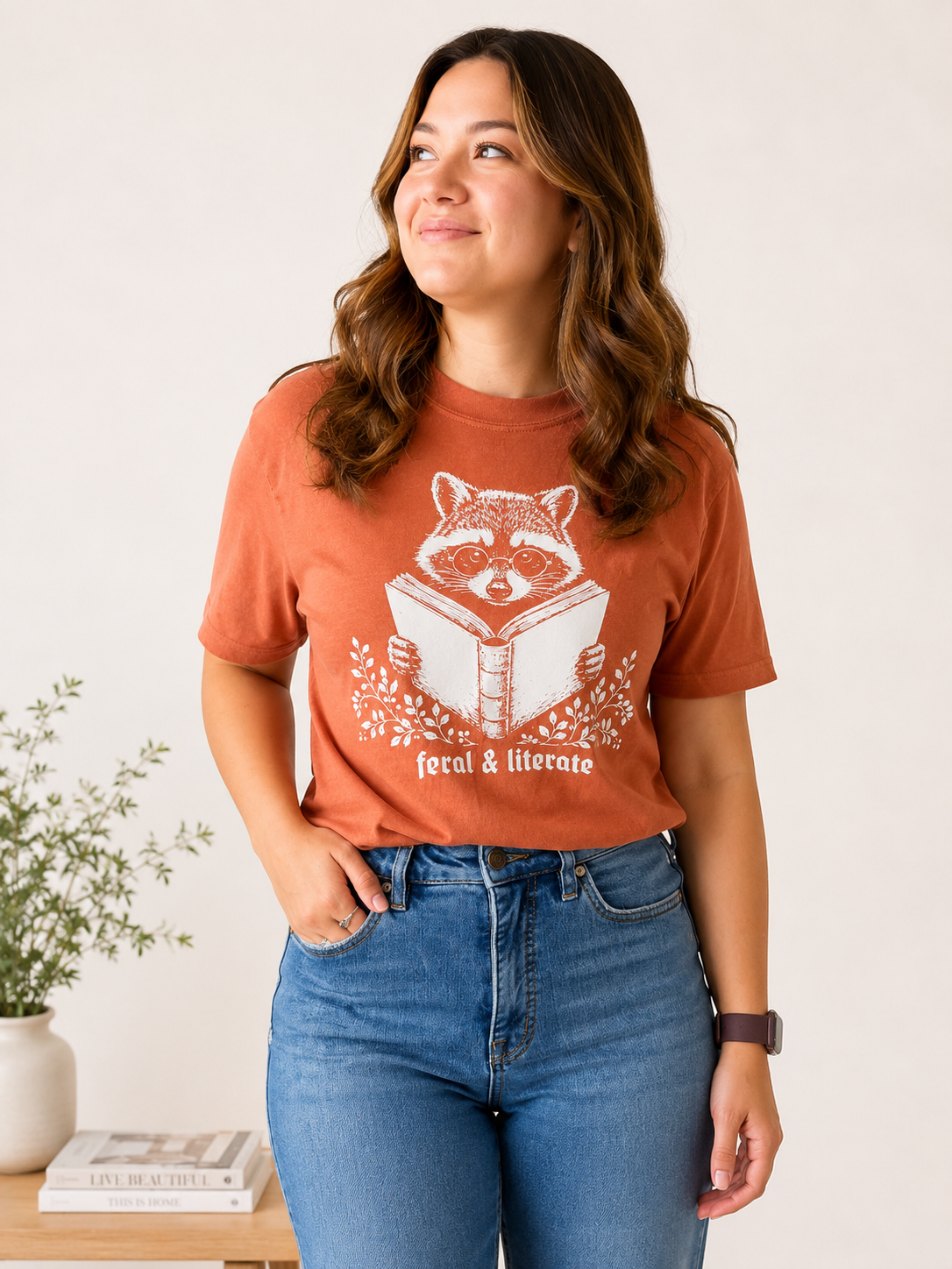A woman wearing a rust-orange graphic t-shirt with a white raccoon reading book design and the text 'fetal & literate', paired with blue jeans, standing against a light beige wall.