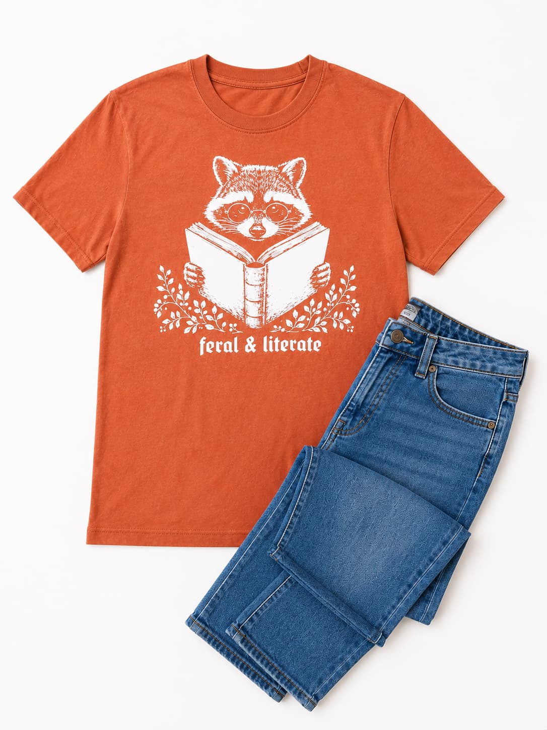 An orange short-sleeve crewneck t-shirt with a white graphic of a raccoon reading a book and the text 'feral & literate', styled flat-lay next to a pair of folded blue jeans on a white background.