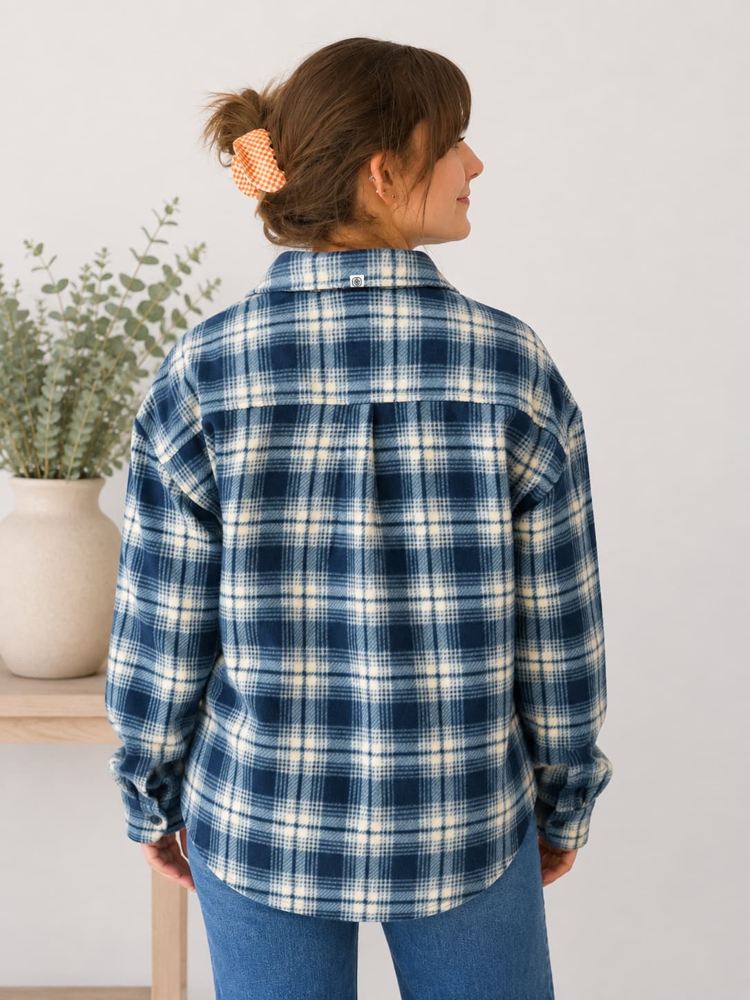 A woman shown from the back wearing a blue and white plaid flannel shirt, paired with blue jeans, standing in a brightly lit indoor setting.