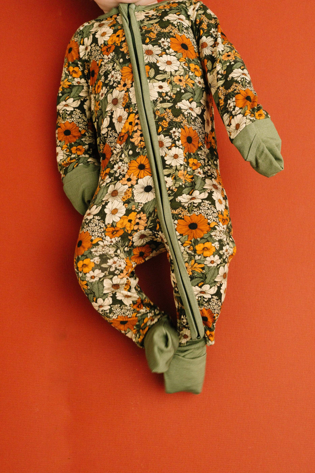A baby footed sleeper with a green, orange, and white floral pattern, displayed on a solid orange-red background.