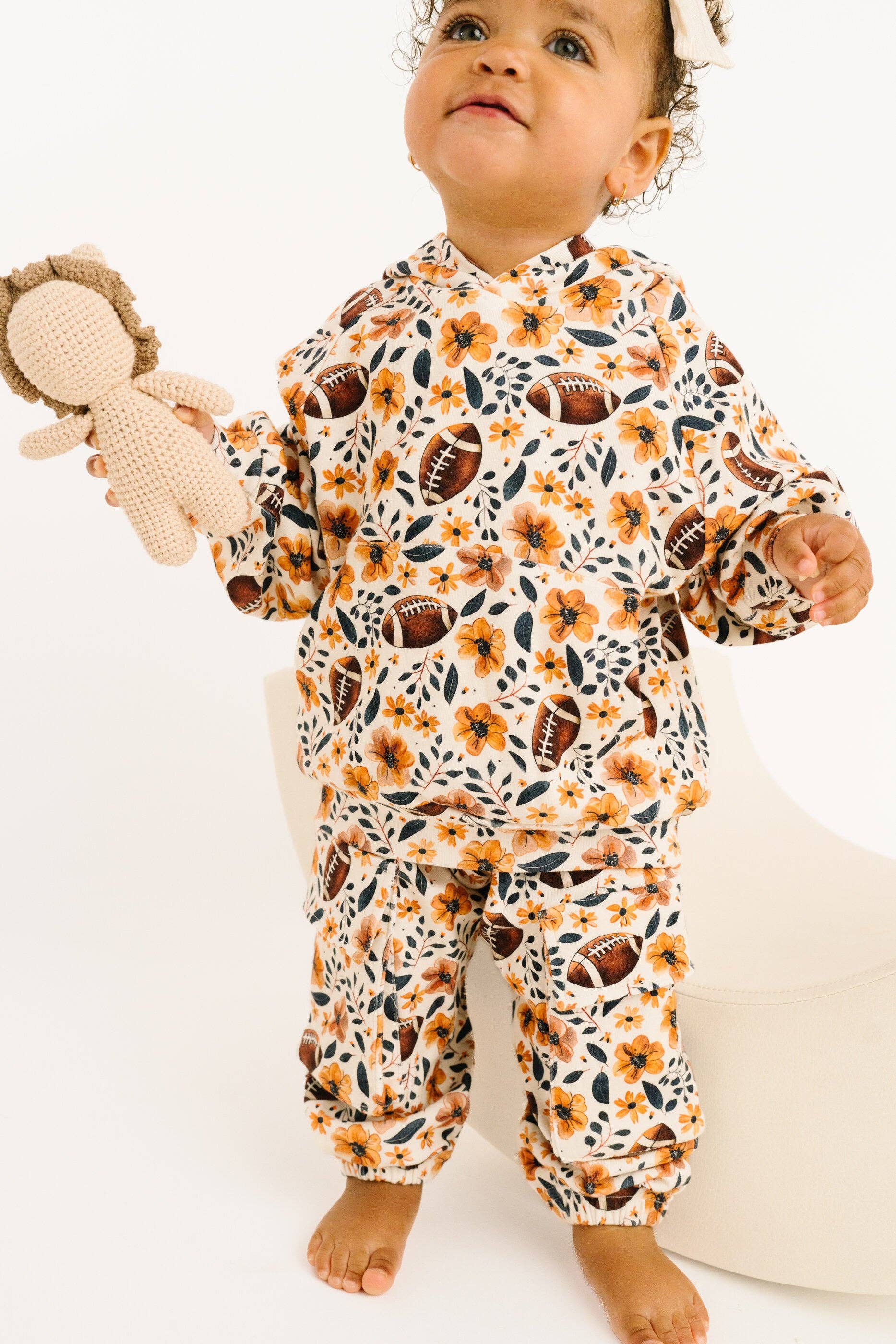 A baby wearing a matching two-piece hooded sweatshirt and pants set with a brown, orange, and black floral and football print, standing barefoot on a white background and holding a small stuffed doll.