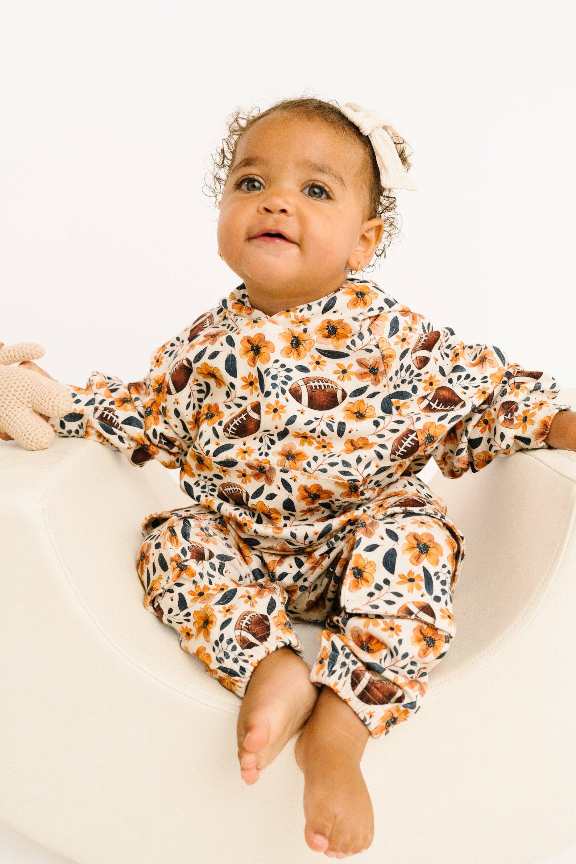 A baby sitting in a white tub wearing a long-sleeve matching pajama set with a floral and football print, along with a white bow headband.