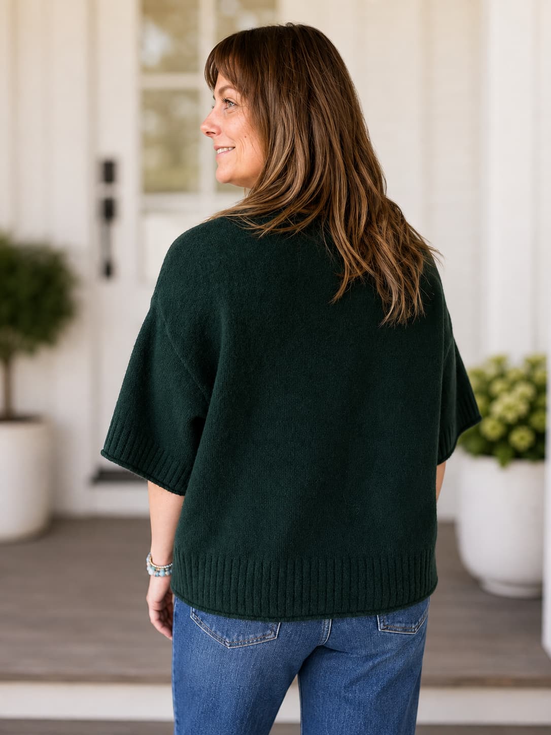 A woman wearing a dark forest green knit sweater and blue jeans, photographed from the back in a bright, modern porch setting.