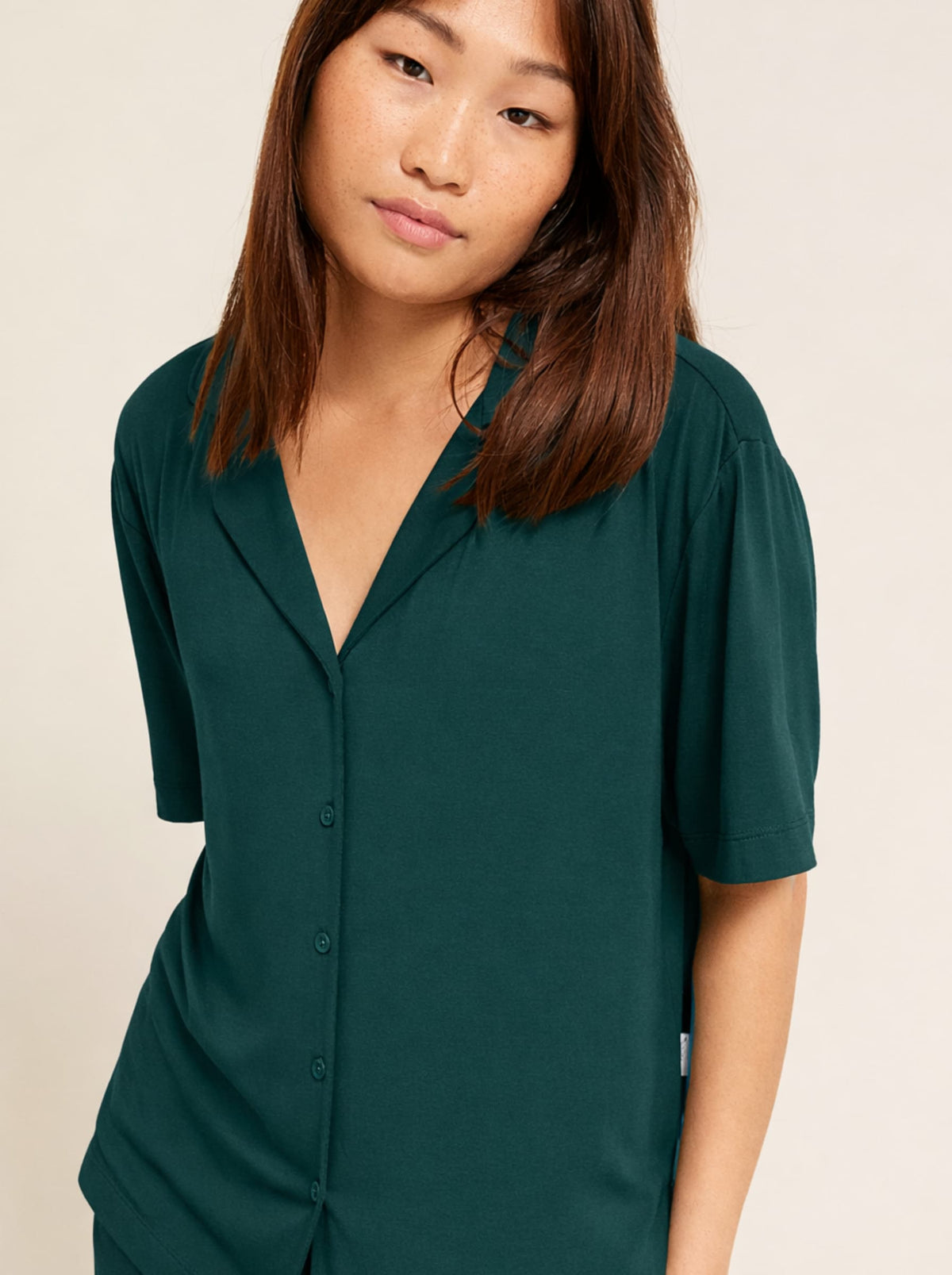 A woman wearing a dark green short-sleeve button-front top with a V-neck and collar, photographed against a plain light beige studio background.