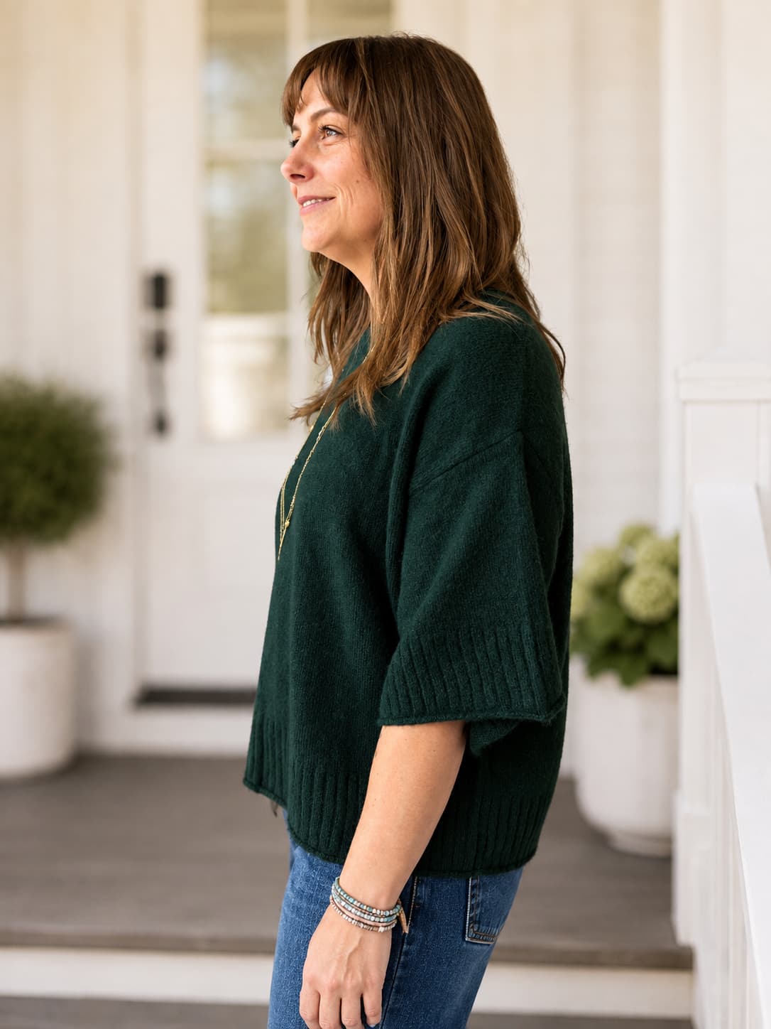 A woman wearing a super soft dark forest green knit sweater and blue jeans, standing on a porch with a white door and potted plants in the background.