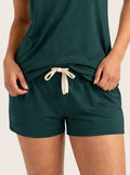 A close-up of a person wearing dark teal green lounge shorts with a cream drawstring and a matching dark teal top, shown against a plain light background.