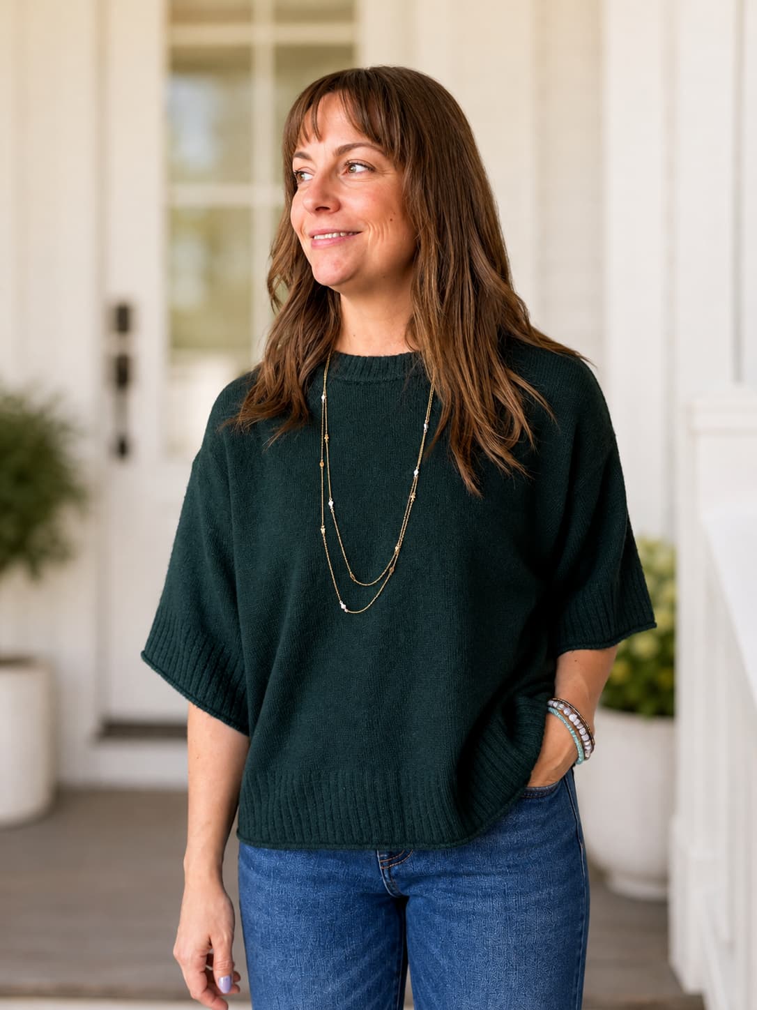 A woman wearing a dark forest green short-sleeve knit sweater with a long gold necklace, beaded bracelets and blue jeans, standing on a porch with a blurred white door and plants in the background.