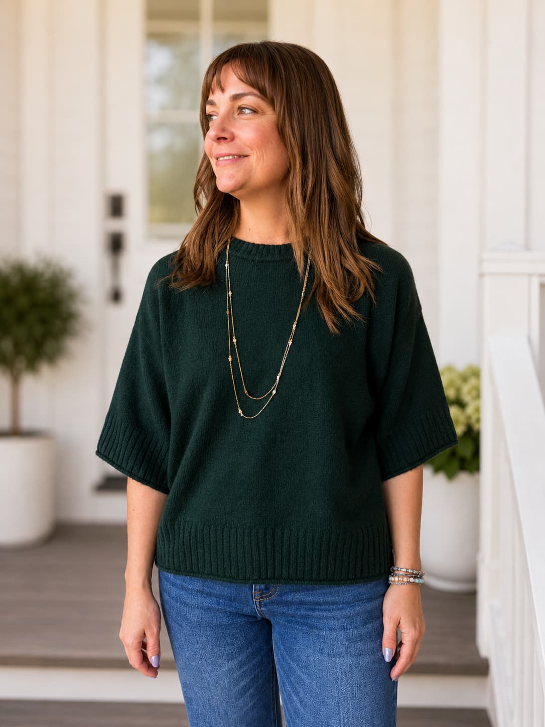 A woman wearing a dark forest green short-sleeve knit sweater and blue jeans, standing on a white porch with potted plants in the background.
