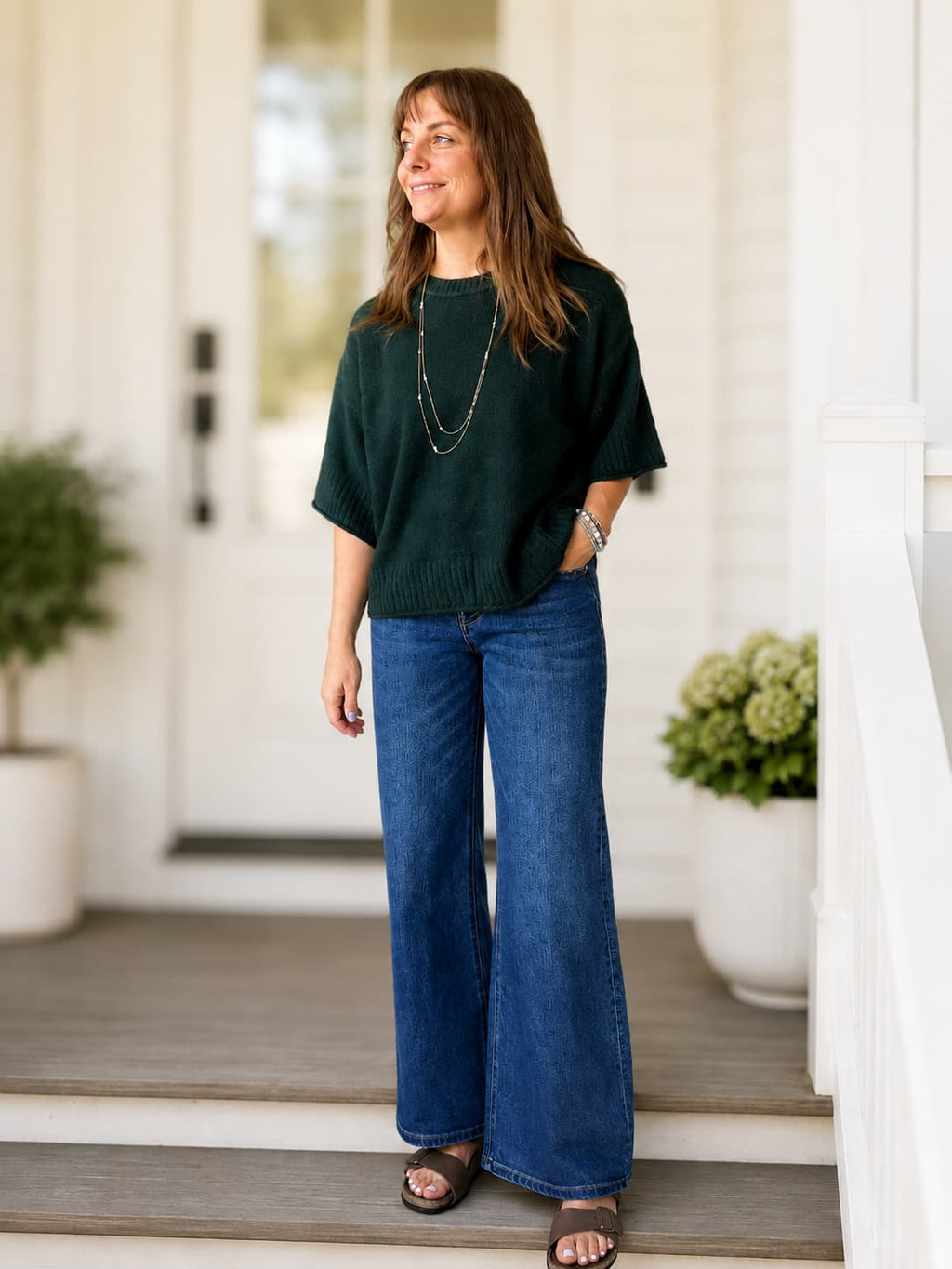A woman stands on a porch wearing a forest green short-sleeve knit sweater, blue wide-leg jeans, and brown sandals.