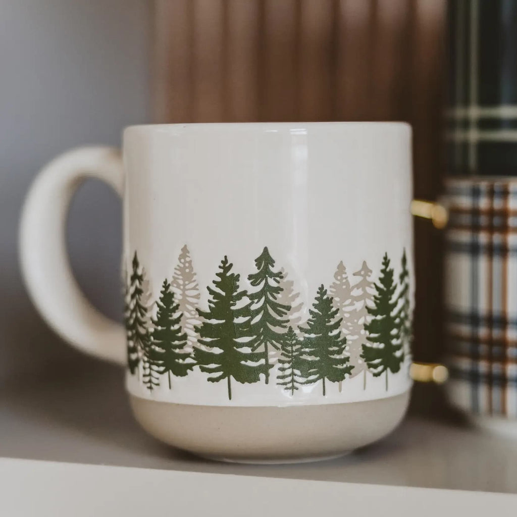 A white ceramic mug with a forest of pine trees printed around the middle, featuring a matte beige base and a gold-toned metal band near the rim. The mug is shown on a shelf with a blurred background.