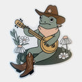 A die-cut sticker featuring a cartoon green frog wearing a brown cowboy hat and boots, playing a mandolin surrounded by white daisies on a light gray background.