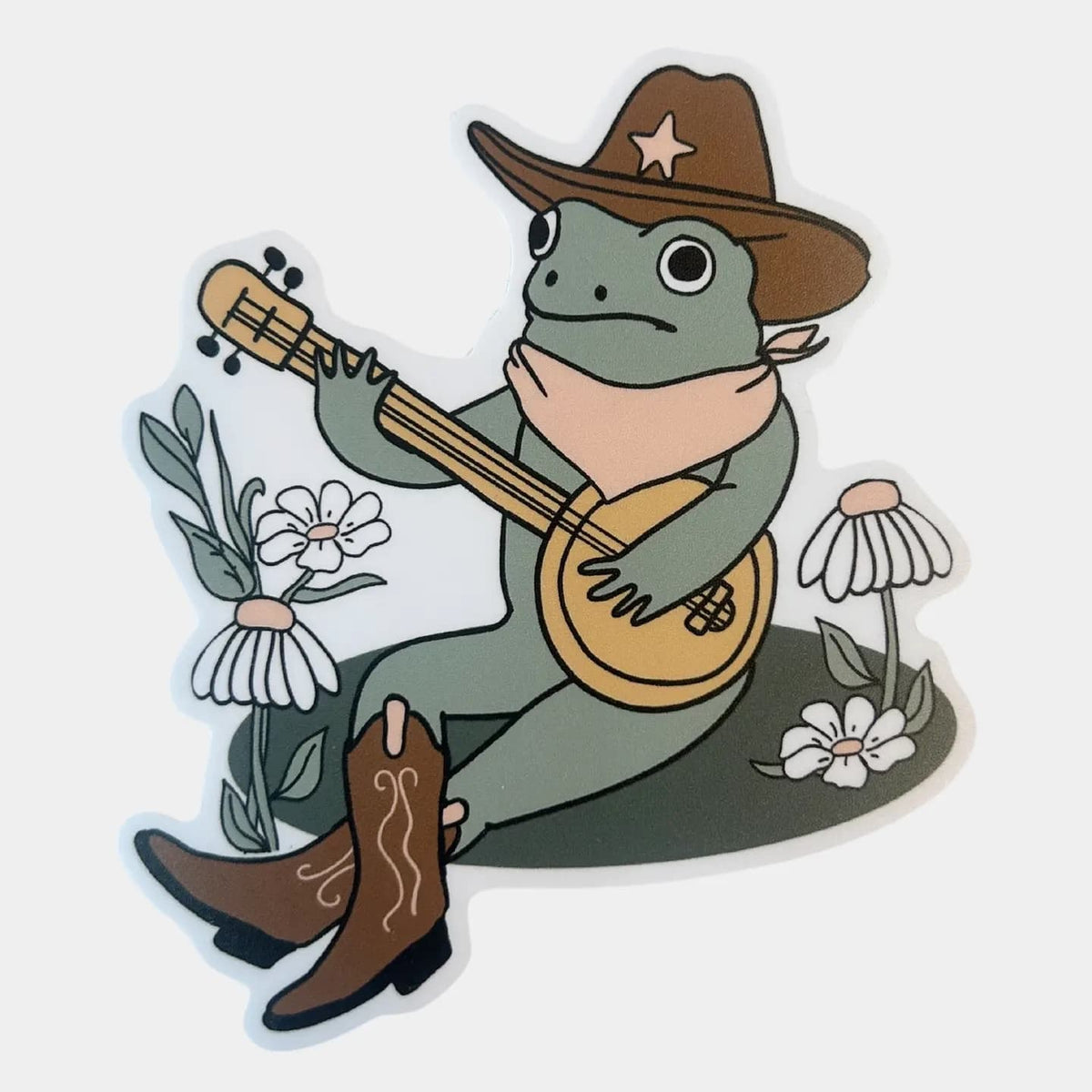 A die-cut sticker featuring a cartoon green frog wearing a brown cowboy hat and boots, playing a mandolin surrounded by white daisies on a light gray background.