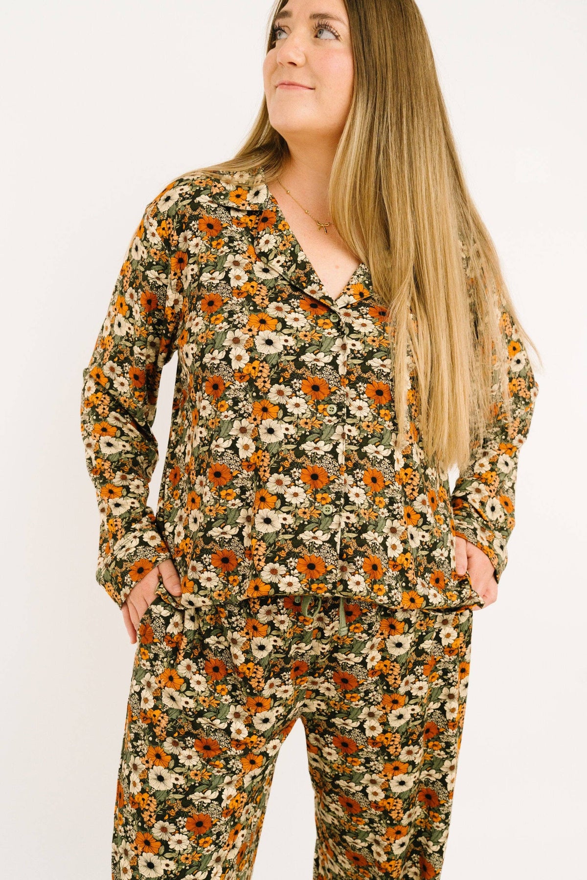 A woman wearing a matching long-sleeve pajama set with a dense floral print in orange, white, and green on a dark background, posed against a plain white studio background.