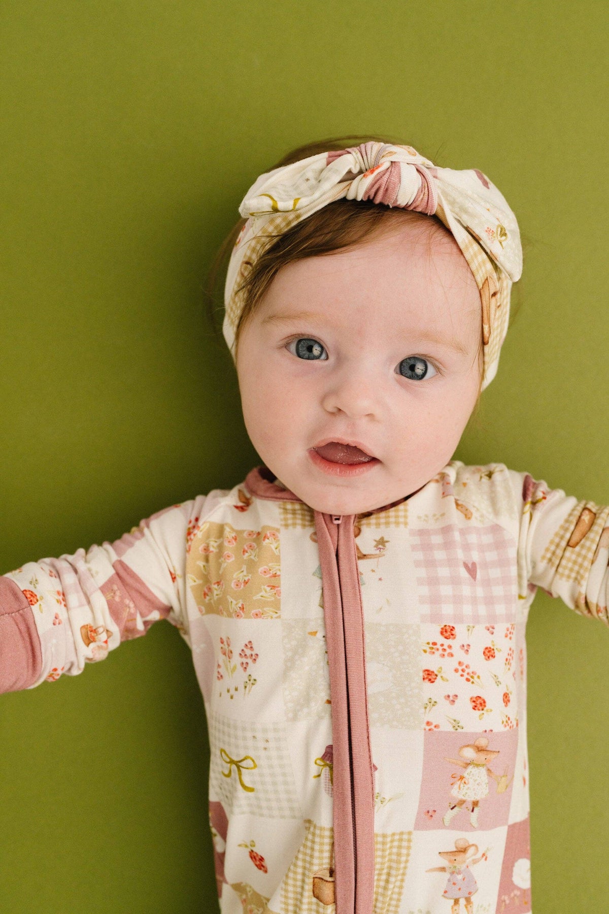 A baby wearing a cream and pink patchwork zip-up onesie and a matching patterned headband, standing against a solid olive green background.