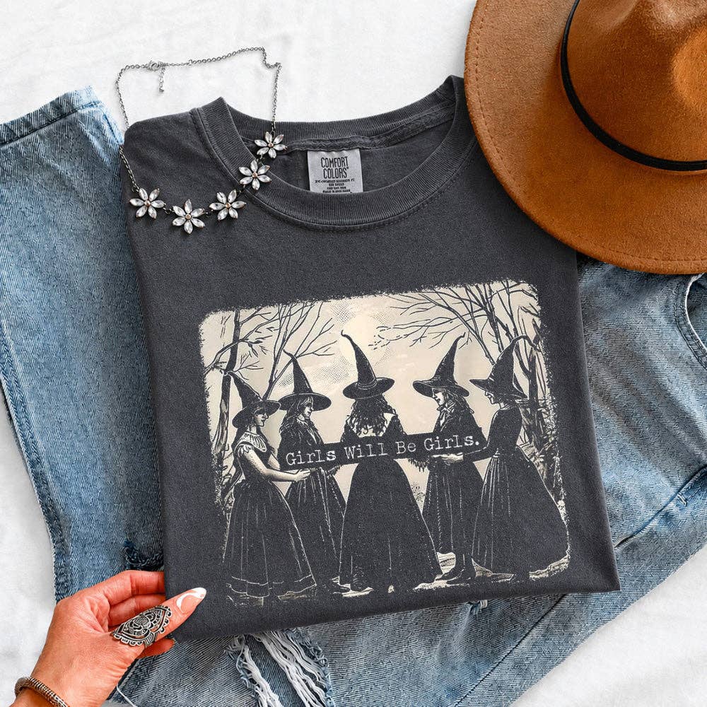 A dark charcoal gray graphic t-shirt featuring a black and white illustration of five witches holding a banner that reads 'Girls Will Be Girls'. The t-shirt is styled in a flat-lay arrangement with light-wash blue jeans, a brown wide-brim hat, and a silver floral necklace, held by a hand at the bottom left.