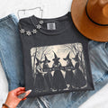 A dark charcoal gray graphic t-shirt featuring a black and white illustration of five witches holding a banner that reads 'Girls Will Be Girls'. The t-shirt is styled in a flat-lay arrangement with light-wash blue jeans, a brown wide-brim hat, and a silver floral necklace, held by a hand at the bottom left.
