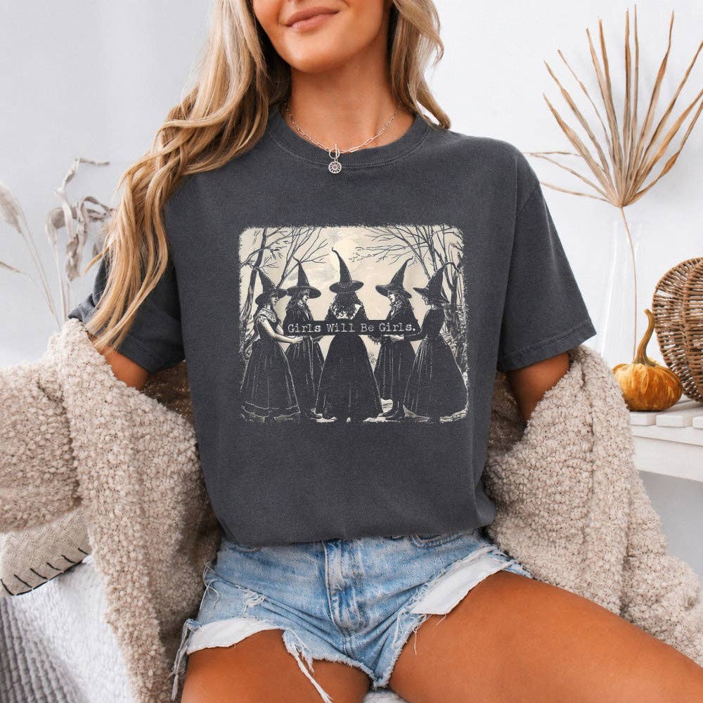 A woman wearing a dark charcoal gray graphic t-shirt with a vintage-style print of four witches and the text 'girls will be witches'. She has a cozy beige sherpa cardigan draped off her shoulders and is wearing denim shorts in a styled indoor setting.