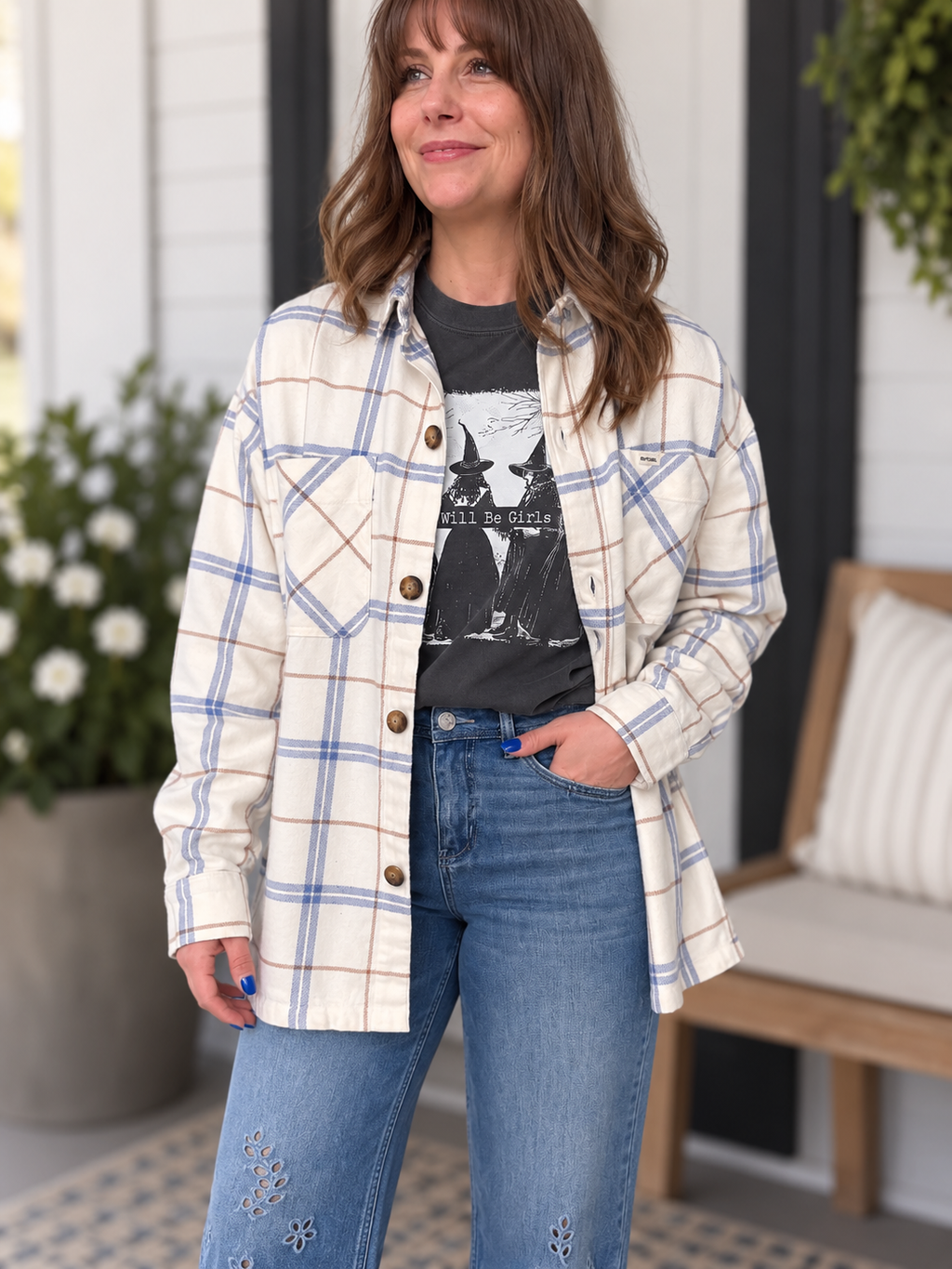A woman wearing a cream and blue plaid button-up shirt over a black graphic t-shirt and blue jeans, standing on a porch.
