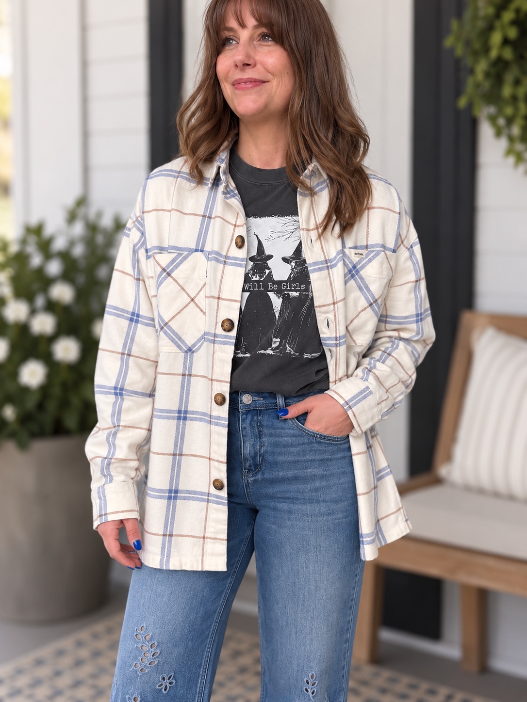 A woman wearing a cream and blue plaid button-up shirt over a black graphic t-shirt and blue jeans, standing on a porch.