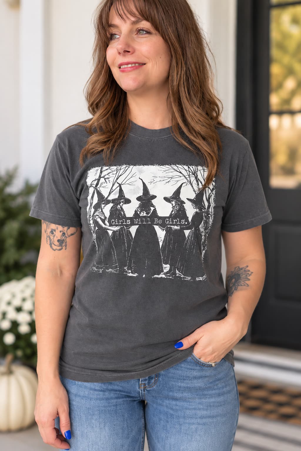 A woman wearing a dark gray graphic t-shirt with a black and white print of witches and the text 'girls will be witches', paired with blue jeans.