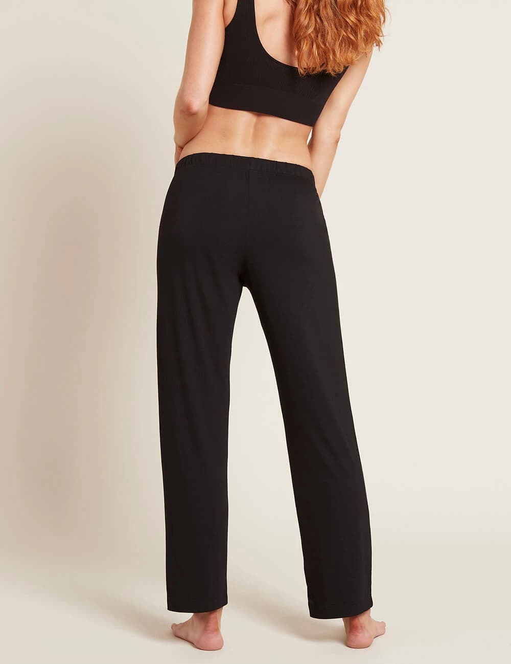 A woman shown from the back wearing black lounge pants and a black sports bra against a plain light beige studio background.