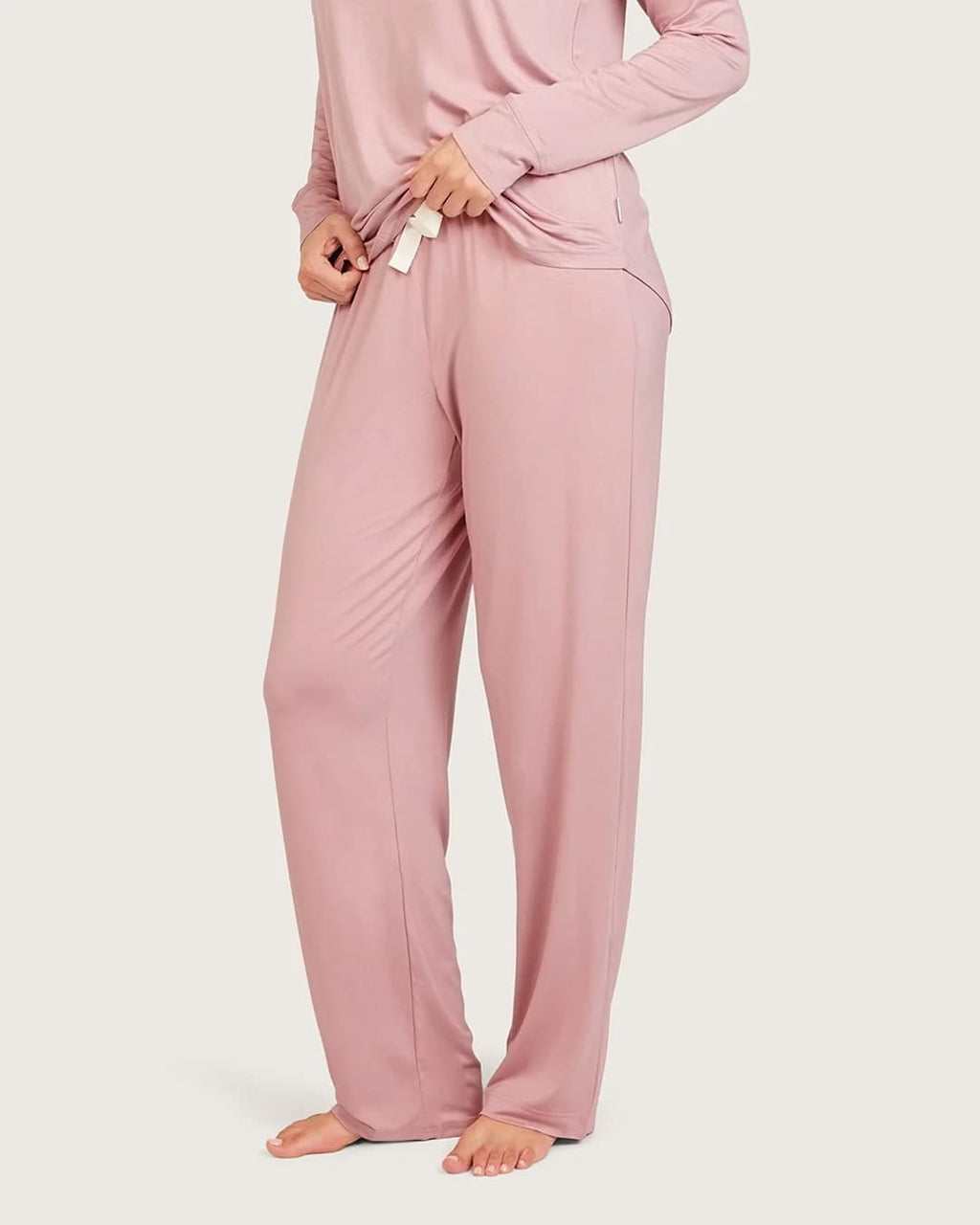 A person wearing a dusty lilac long-sleeve pajama top and matching wide-leg pants, standing barefoot against a plain light beige studio background.