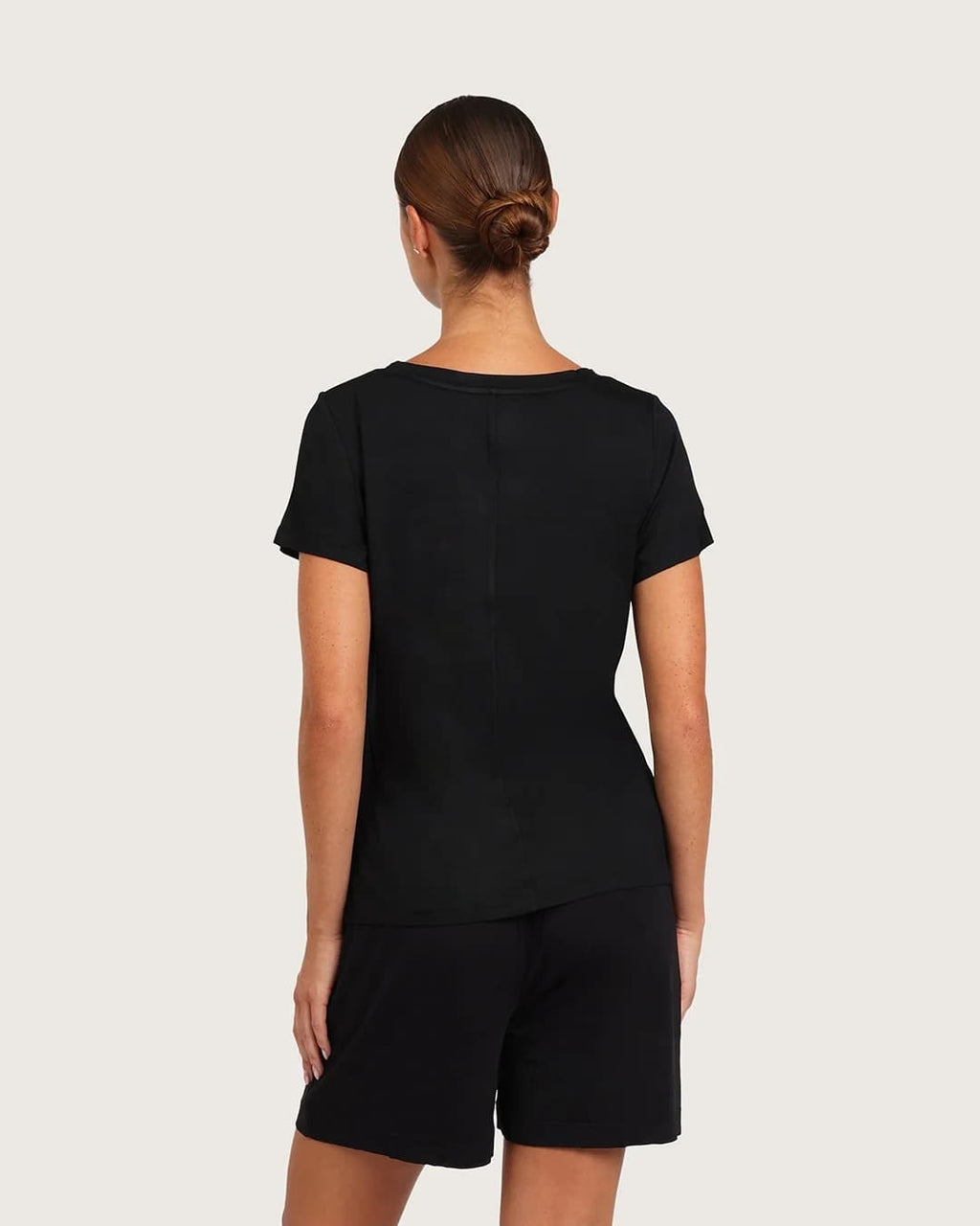 A woman viewed from the back wearing a black short-sleeve t-shirt and matching black shorts against a light gray studio background.