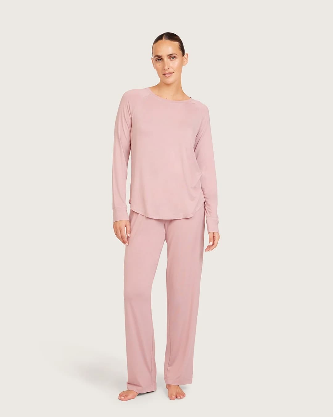 A woman modeling a two-piece dusty lilac loungewear set, consisting of a long-sleeve crewneck top and matching wide-leg pants, against a solid light gray background.