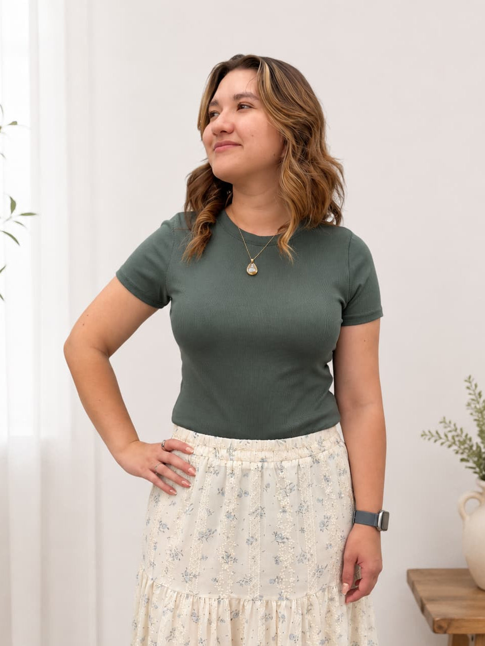 Woman wearing a green top and floral skirt standing against a white wall with plants.