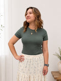 Woman wearing a green top and floral skirt standing against a white wall with plants.