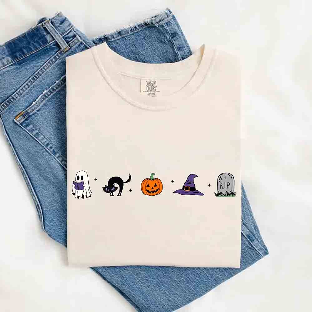 A folded cream-colored t-shirt with a Halloween-themed graphic print, laid flat on top of blue jeans against a white background. The graphic features a ghost, a black cat, a pumpkin, a witch hat, and a tombstone.