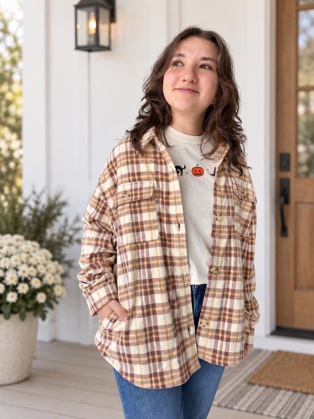 A young woman stands on a front porch wearing an oversized brown and cream plaid flannel shirt over a white graphic t-shirt and blue jeans.