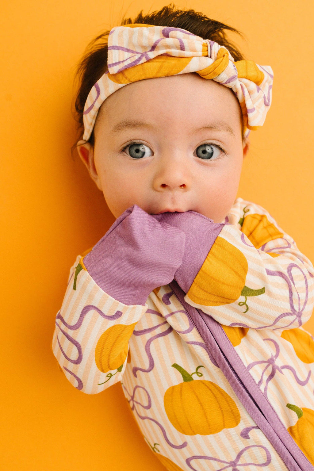 A baby wearing a cozy Halloween-themed zip-up sleeper and matching headband, featuring a pumpkin print on a cream background with purple swirls and a purple zipper, posed against a solid orange background.