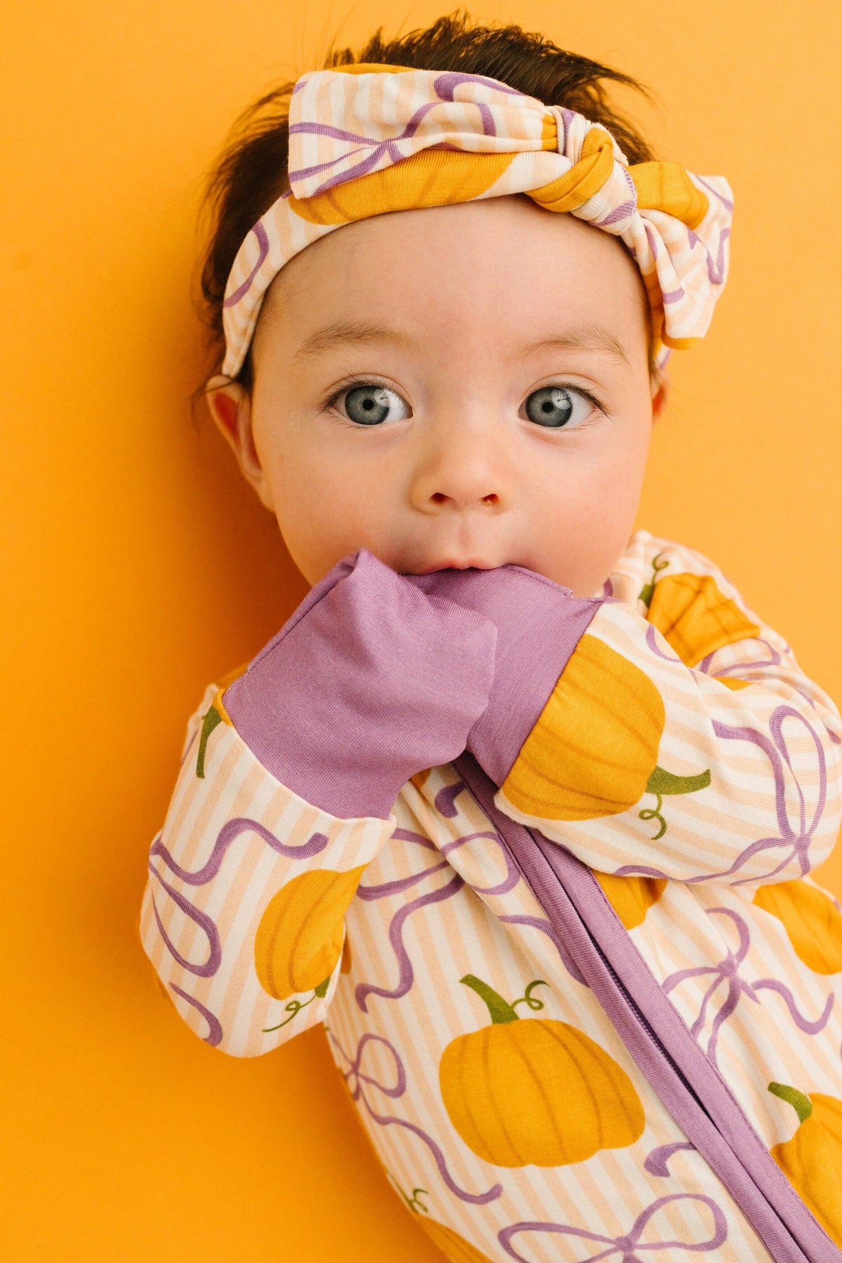 A baby wearing a cozy Halloween-themed zip-up sleeper and matching headband, featuring a pumpkin print on a cream background with purple swirls and a purple zipper, posed against a solid orange background.