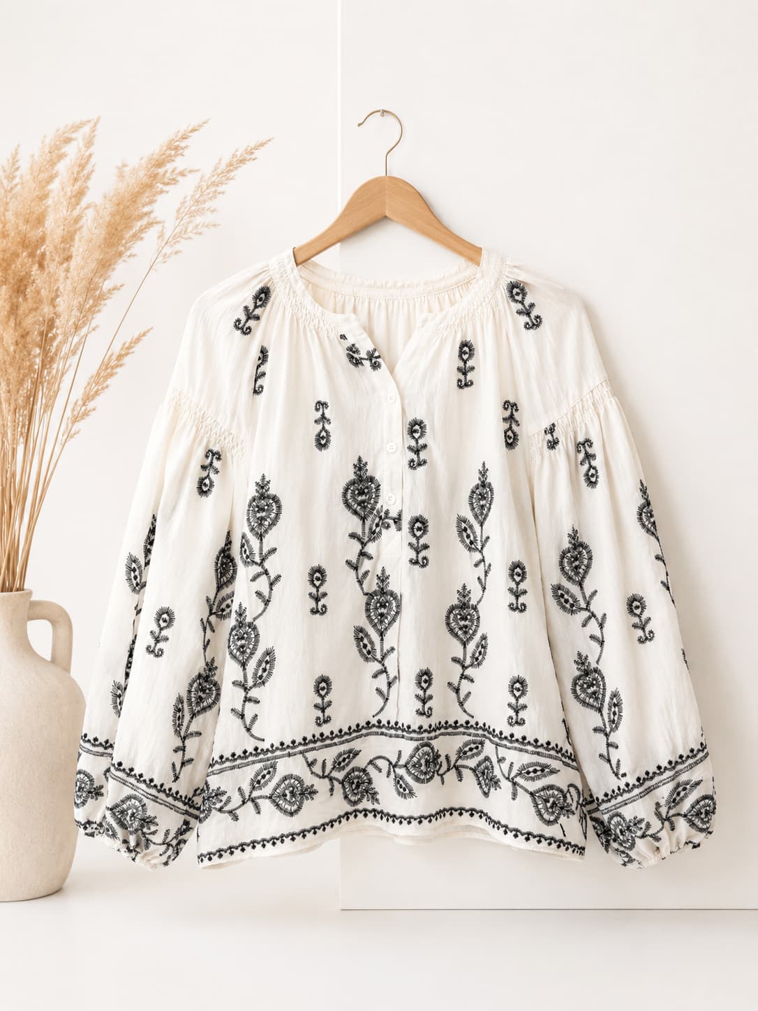 White blouse with black floral embroidery on a wooden hanger against a white background.