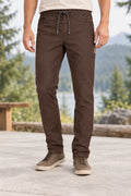 A person wearing dark brown slim-fit drawstring pants, standing outdoors on a concrete patio with a blurred forest and mountain background.