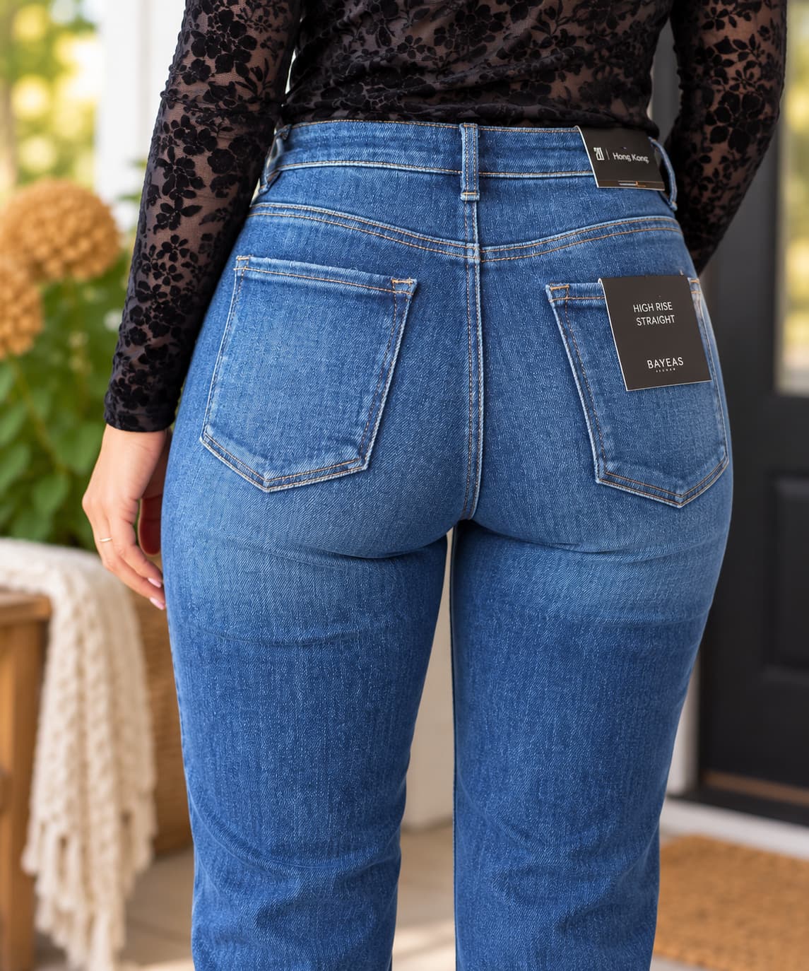 Rear view of a person wearing blue high-rise jeans with a black lace long-sleeve top, showing the back pockets and a Bayes brand patch on the jeans.