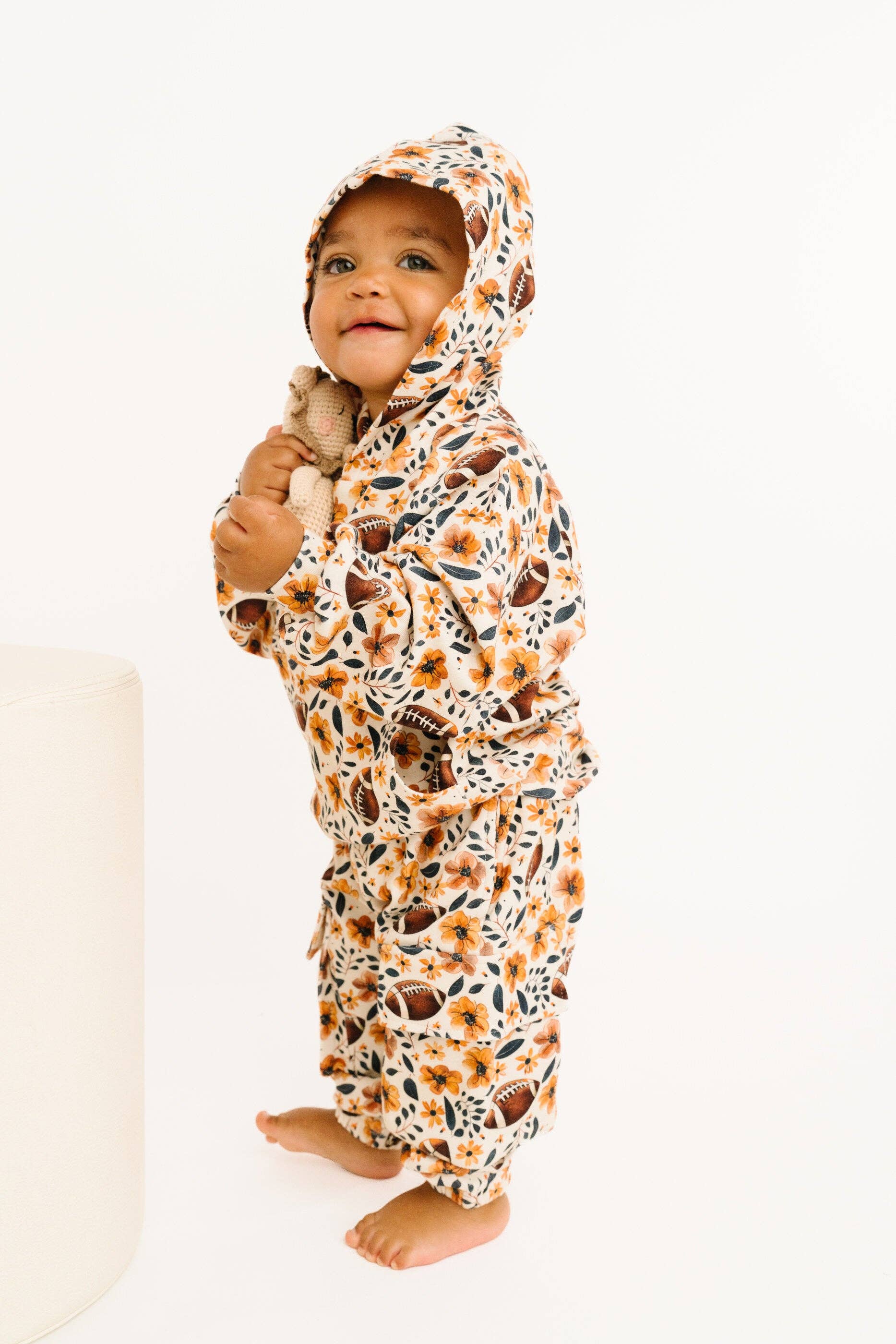 A baby wearing a hoodie and cargo pant set with brown, black, and orange floral pattern, standing against a plain white background.