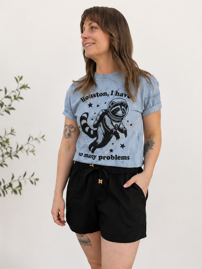 Woman wearing a blue t-shirt with a graphic design and text, standing in a room with a plant and rug.
