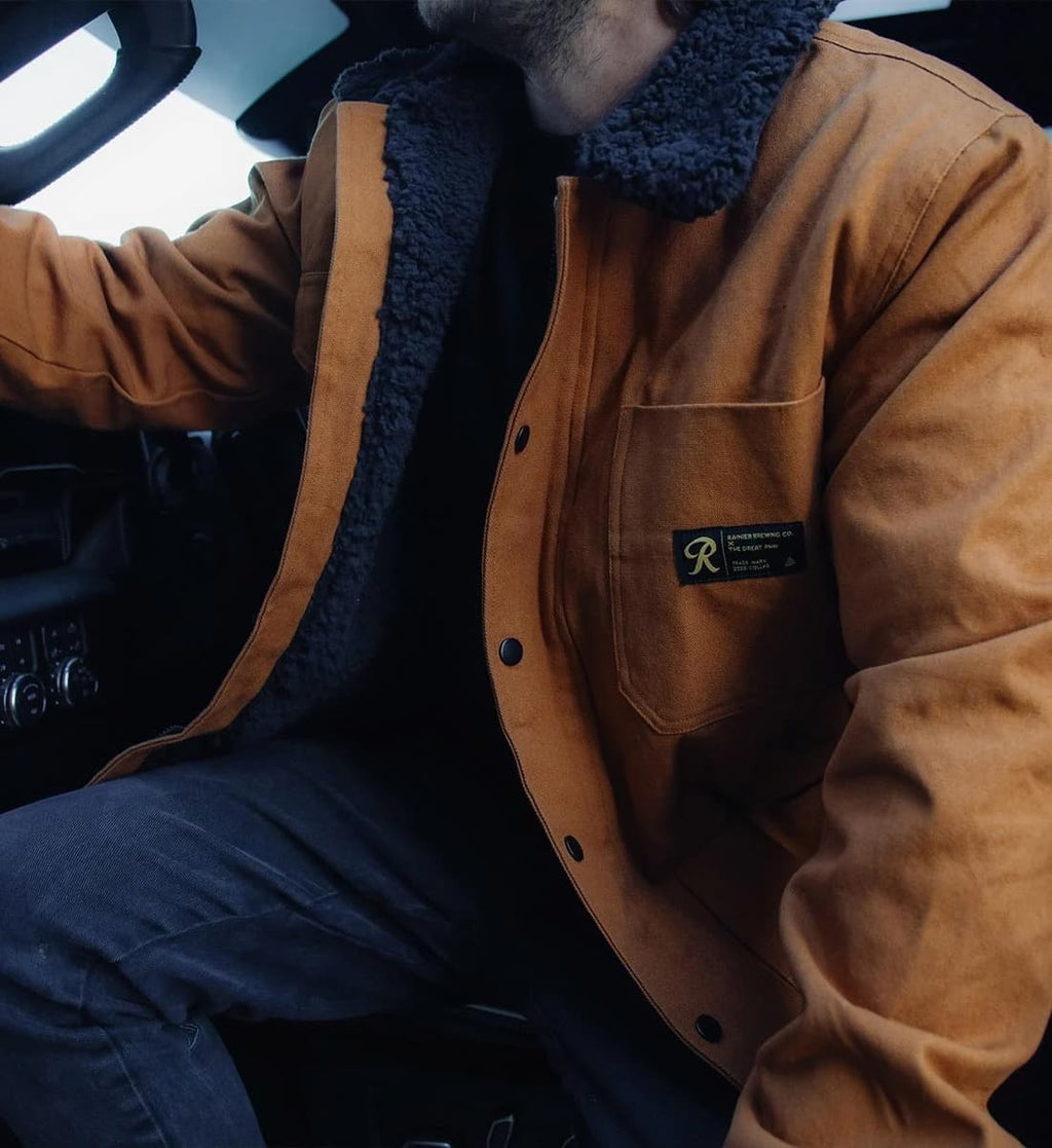 A person sitting inside a car wearing a tan canvas jacket with a dark navy fleece collar over a black shirt and dark pants. A black brand patch is visible on the chest pocket.