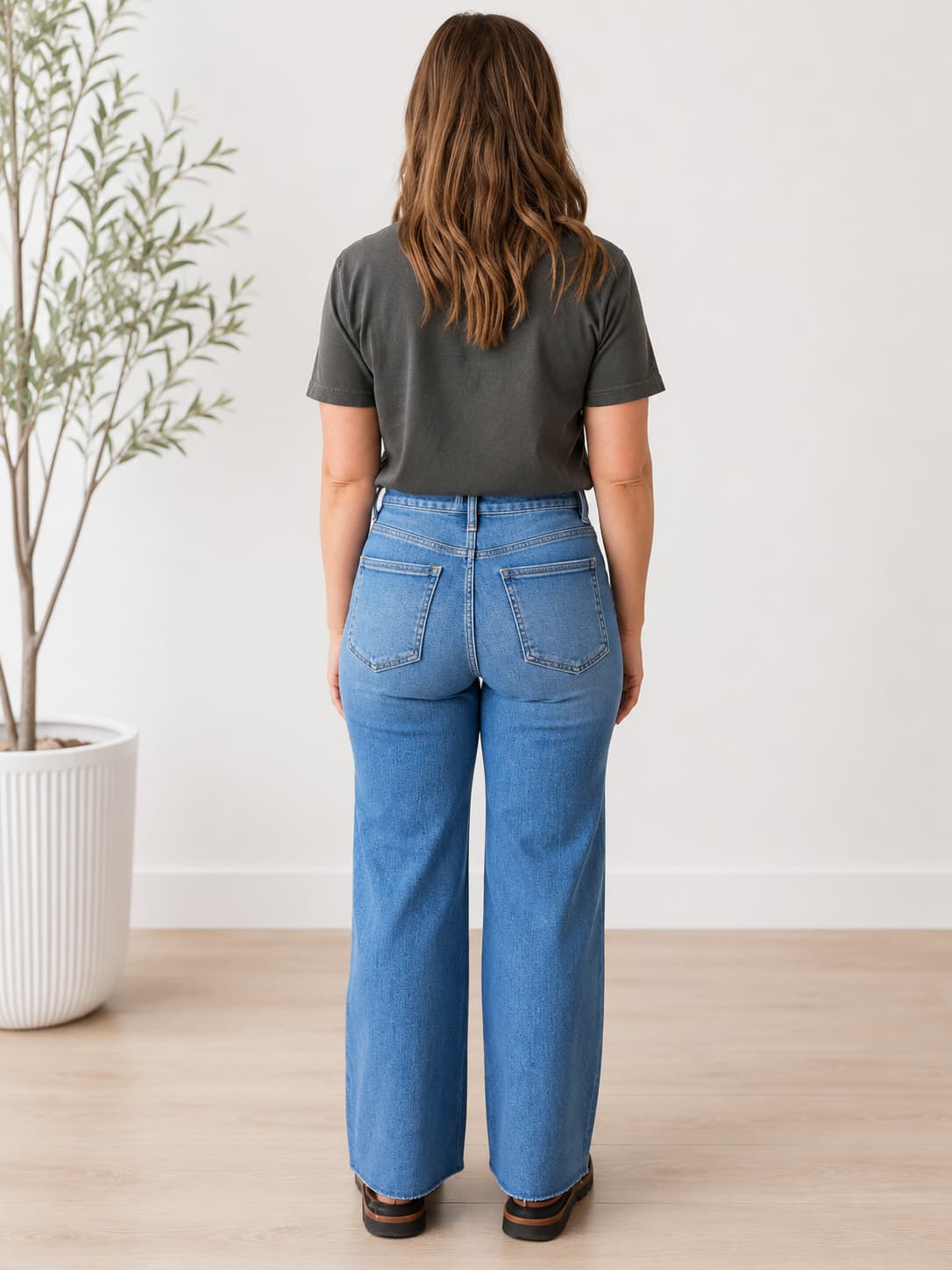 A woman viewed from behind wearing high-waisted, wide-leg blue jeans and a dark gray t-shirt, standing in a bright room with a light wood floor and a potted plant on the left.