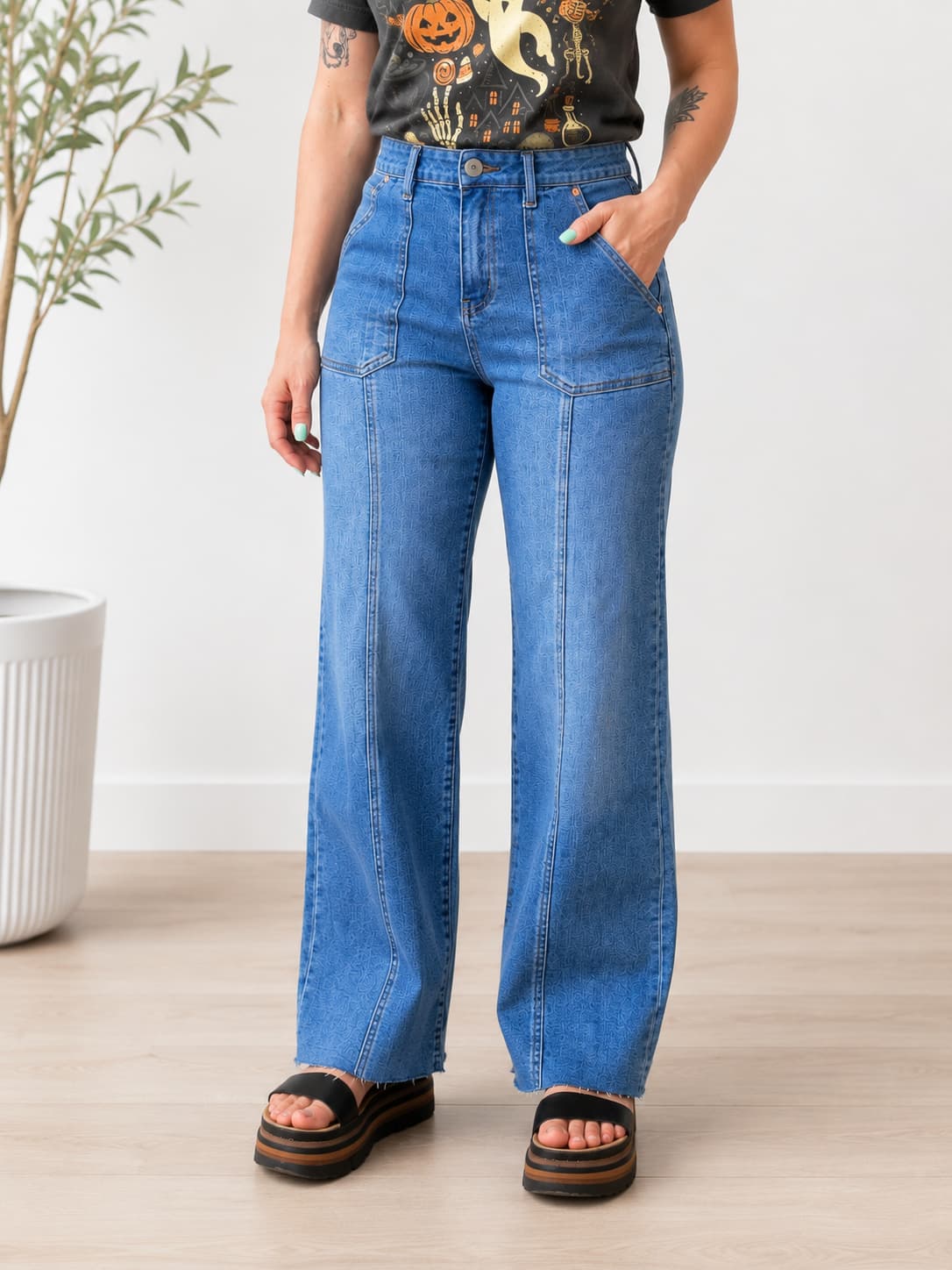 A person wearing high-waisted, wide-leg blue denim jeans with a black graphic t-shirt and black platform sandals, standing in a brightly lit room with a light wood floor and a white wall.